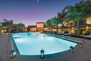Piscina con sillas de descanso y palmeras, rodeada de edificios con grandes ventanales e iluminación exterior.