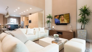 Ein modernes Wohnzimmer mit einer Couch, einem Couchtisch und einem Fernseher. Im Hintergrund befinden sich Pflanzen und eine Küche.