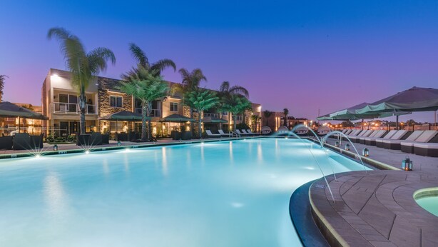 Piscina com espreguiçadeiras e guarda-sóis, rodeada por edifícios com varandas e palmeiras.