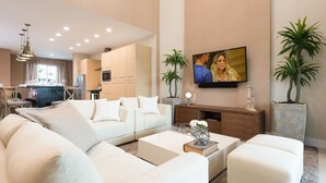 Una sala moderna con sofá, mesa de centro y televisor. Se ven plantas y una cocina al fondo.
