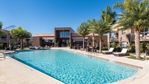 A swimming pool with lounge chairs and palm trees, surrounded by buildings with large windows and a clear sky. - Gorgeous Home/Near Disney/Parking/BBQ/Free Wi-Fi/ Heated Shared Pool (Kissimmee)