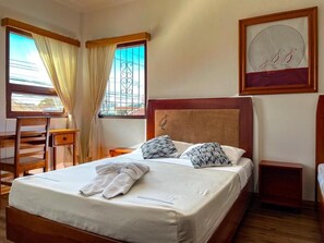 A neatly made bed with a white comforter and two patterned pillows, a wooden nightstand with a lamp, a wooden chair, and a desk with a chair. - Luz De Luna Hotel Barrio Escalante (San José)
