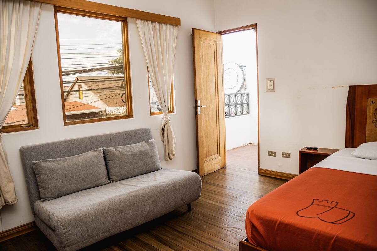 Ein Schlafzimmer mit grauem Sofa, einem Bett mit orangefarbener Bettdecke, Holzboden und einer Holztür, die auf einen Balkon führt.