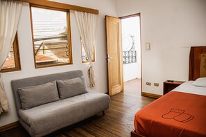 Ein Schlafzimmer mit grauem Sofa, einem Bett mit orangefarbener Bettdecke, Holzboden und einer Holztür, die auf einen Balkon führt.