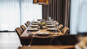 Un comedor con una mesa larga dispuesta para una comida, rodeada de sillas, con una lámpara encima y cortinas en las ventanas.