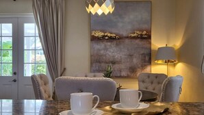 Zona pranzo con tavolo in marmo, due tazze e piattini bianchi, vassoio dorato, poltrona trapuntata, tavolino con lampada e un quadro appeso al muro.