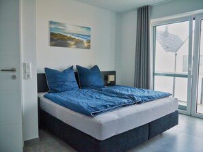 A modern bedroom with a large bed, a nightstand, a wall-mounted lamp, and a window with curtains.