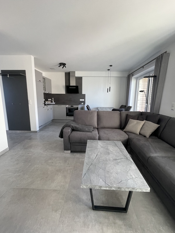 A modern living room with a grey sectional sofa, a marble coffee table, and a kitchen in the background.