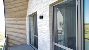 A modern building with a wooden slatted exterior, large sliding glass doors, and a metal balcony railing. - Vacation cottage on the dike (Büsum)