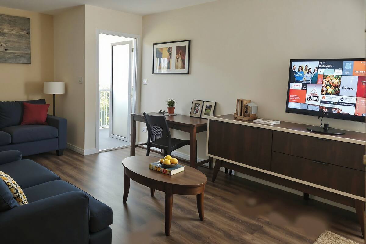 A living room with a sofa, a coffee table with books and fruit, a TV, and a dining area with a chair and table.