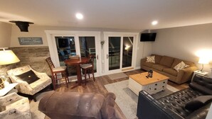 A living room with a brown leather sofa, a wooden coffee table, a brown armchair, a patterned armchair, a dining table with chairs, and a sliding glass door leading to an outdoor area. - Riverfront Cabin – Summertime Escape in the Shenandoah Valley (Shenandoah)