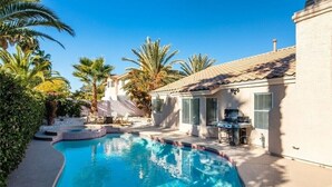 A swimming pool with a curved edge, surrounded by palm trees and a white building with a tiled roof. - Modern and Bright Resort Style Home w/Heated Pool (Las Vegas)