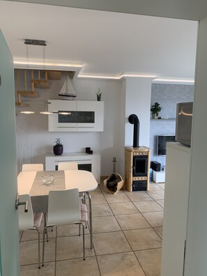 Une cuisine moderne avec des meubles blancs, un four encastré et un poêle en bois. On y trouve une table à manger avec des chaises et un luminaire suspendu.