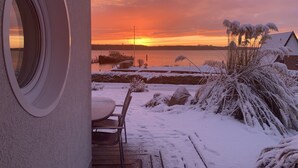 Ein Sonnenuntergang über einem Gewässer, mit einem Steg und einem Boot im Blick. Schnee liegt auf dem Boden und an den Pflanzen.