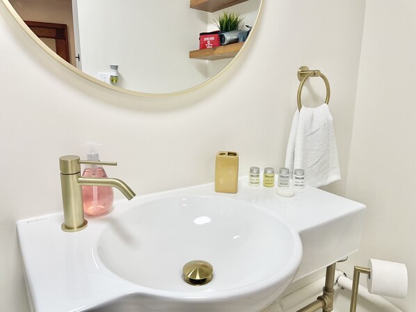 A bathroom with a white sink, gold faucet, and a round mirror. There are toiletries on the sink and a towel hanging on a gold towel ring.