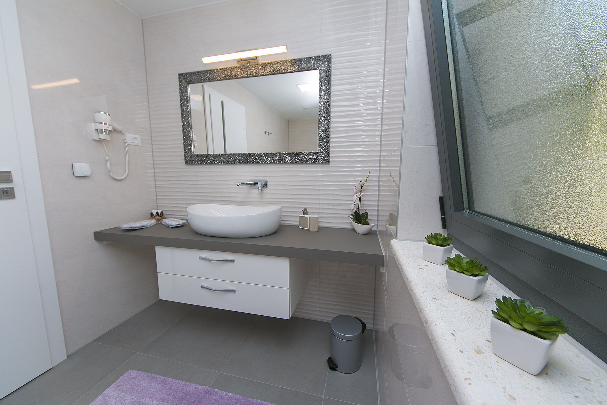 Un baño moderno con un lavabo blanco, un espejo grande y un ventanal con vidrio esmerilado.