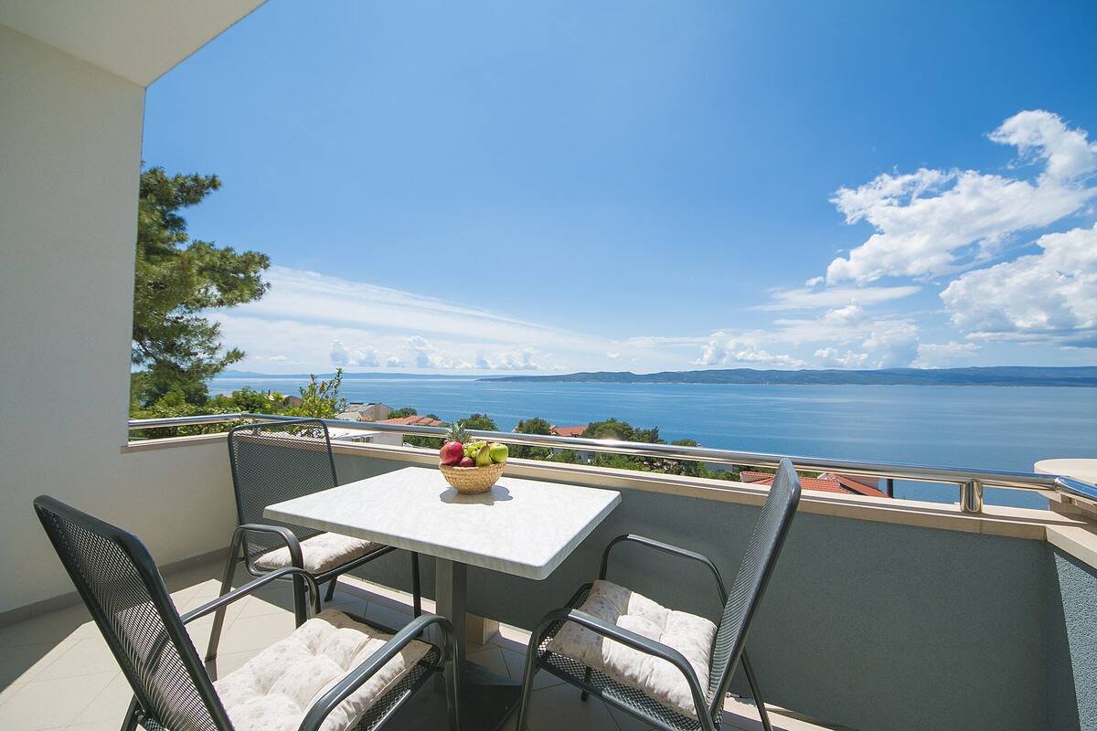 Un balcon avec une table et des chaises, un panier de fruits, et une vue sur la mer et les montagnes.