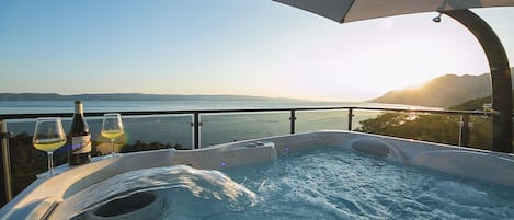Ett bubbelbad med utsikt över havet och bergen, ett bord med vinglas och vinflaskor samt ett parasoll som ger skugga.