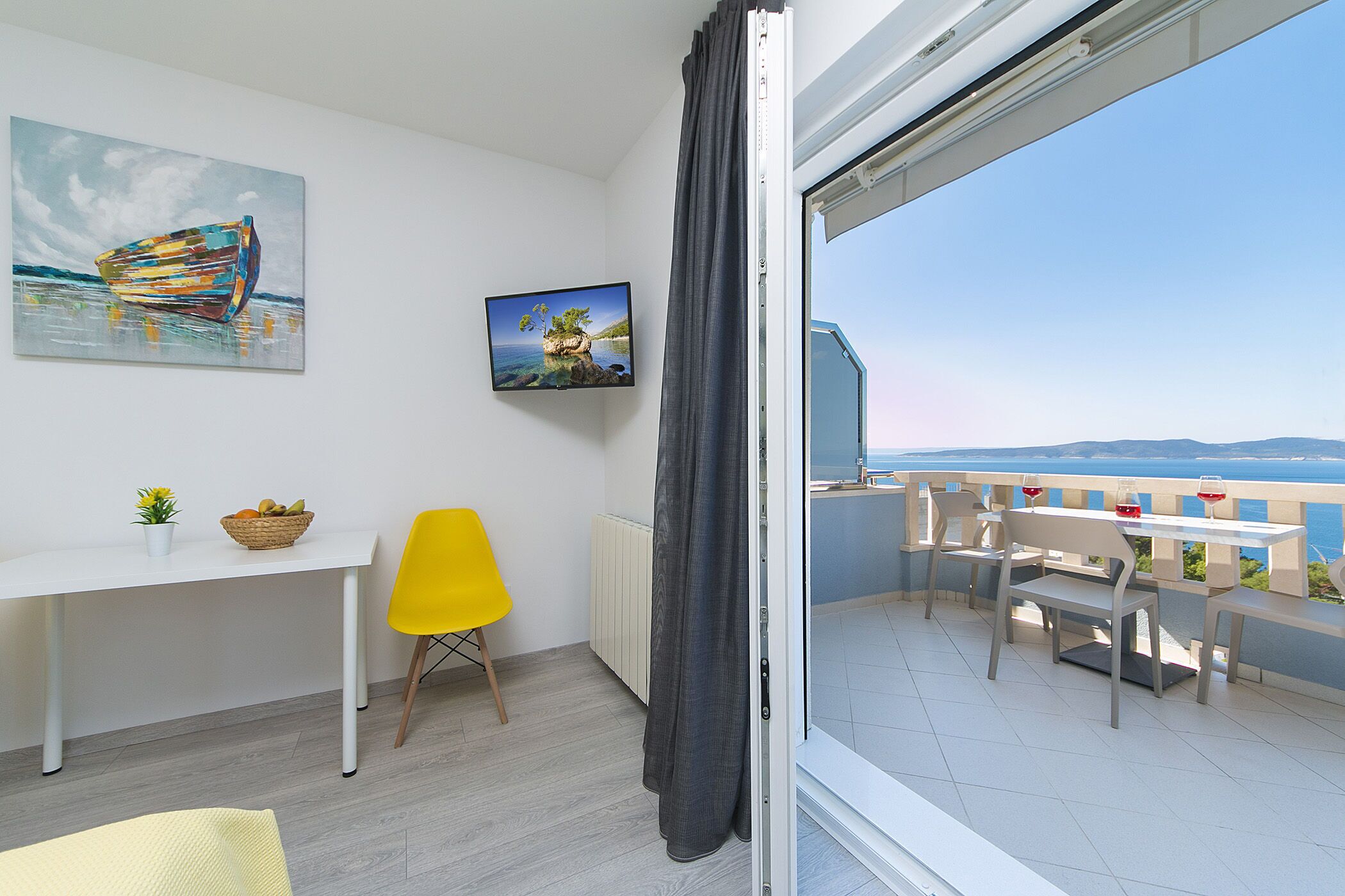 Un balcon avec une table et des chaises, une chaise jaune, un bureau blanc avec un panier, et deux tableaux accrochés au mur.