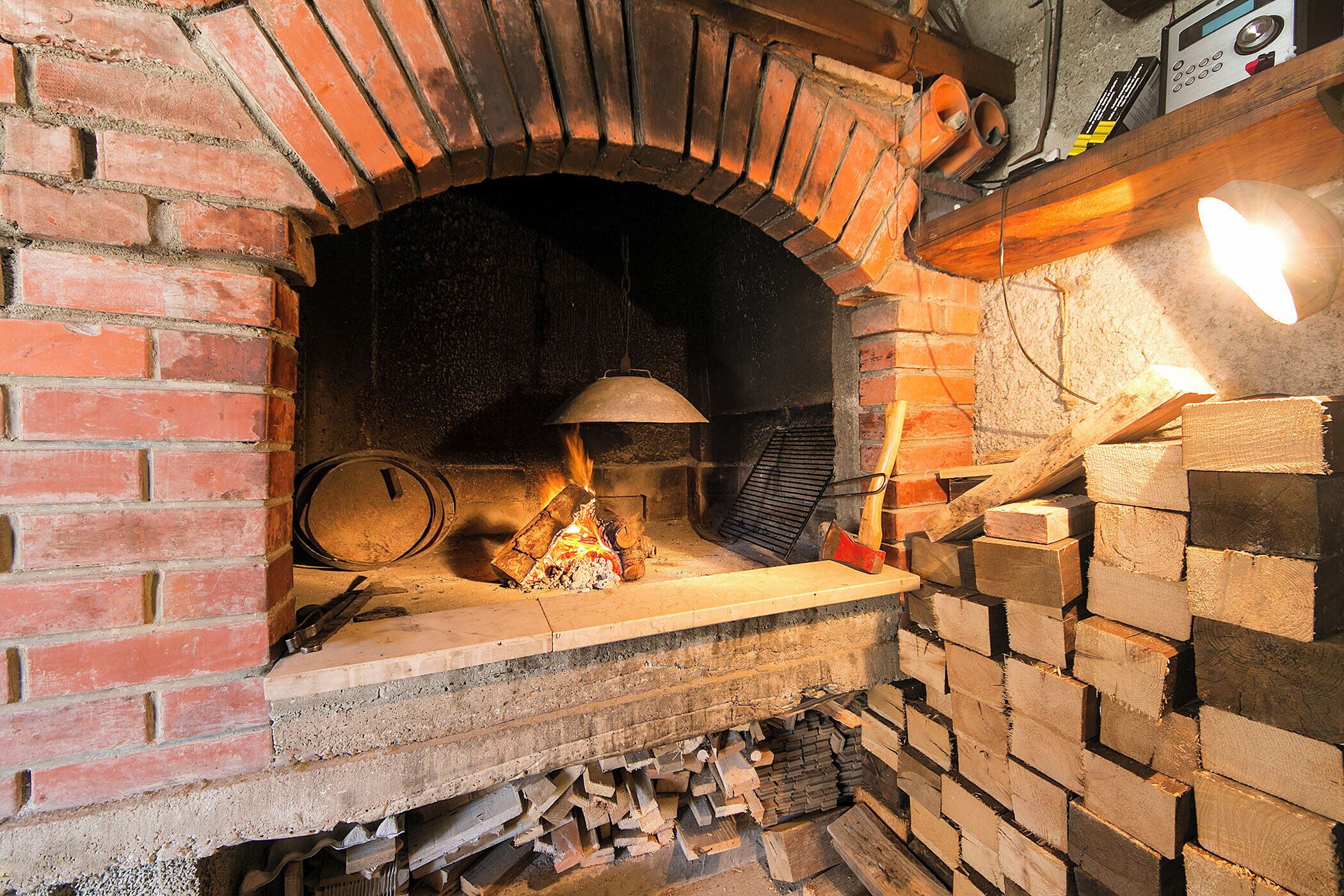 A brick oven with a fire burning inside, surrounded by wood logs and a bell.