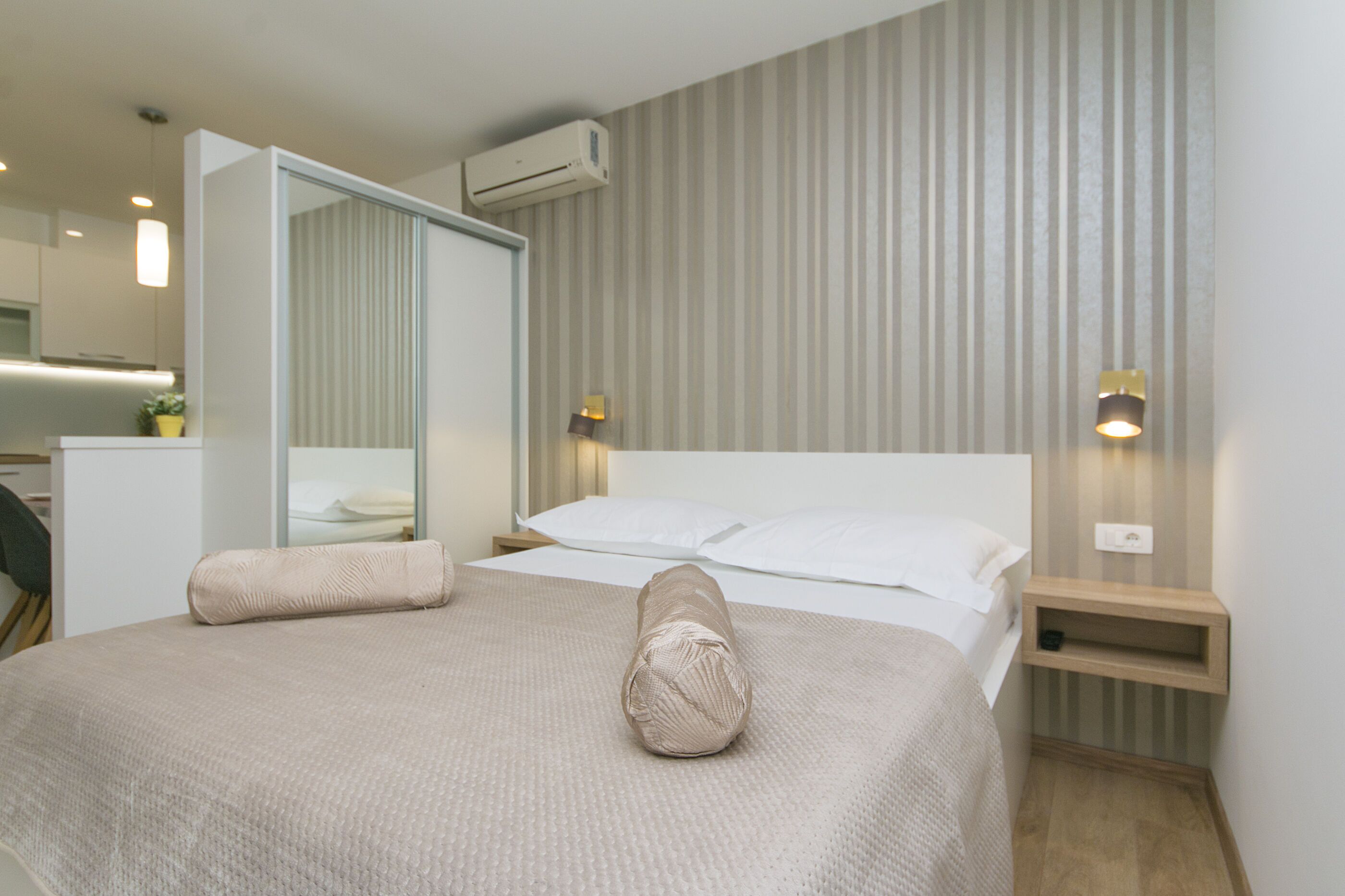 A hotel room with a large bed, a bedside table with a lamp, a mirror, and an air conditioning unit on the wall.