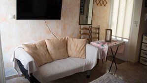 A room with a white sofa, a small table with a pink tablecloth, a wooden chair, a TV mounted on the wall, and a window with blinds. - Cosy and Large Studio in Heraklion Crete, Greece (Heraklion)