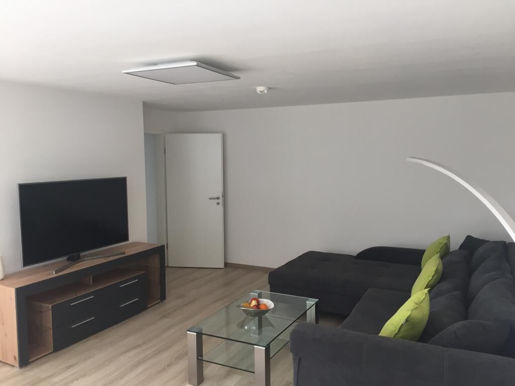 A modern living room with a flat-screen TV on a wooden TV stand, a glass coffee table, a dark sofa, and a door in the background.