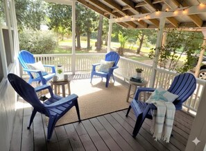 A covered porch with blue chairs and a small table, surrounded by a white fence and trees.