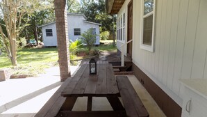 A wooden picnic table with benches under a covered porch, a lantern on the table, and a house in the background. - The Bay Cottage Adventure (Pensacola)