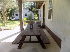 Un tavolo da picnic in legno con panche sotto un portico coperto, una lanterna sul tavolo e una casa sullo sfondo.
