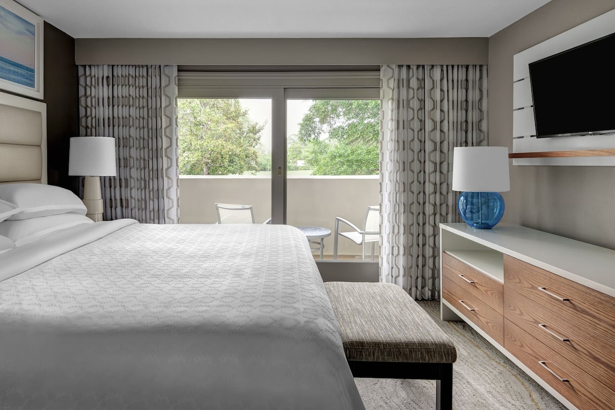 A modern hotel room with a large bed, a balcony with a view, a flat-screen TV, and a wooden dresser.