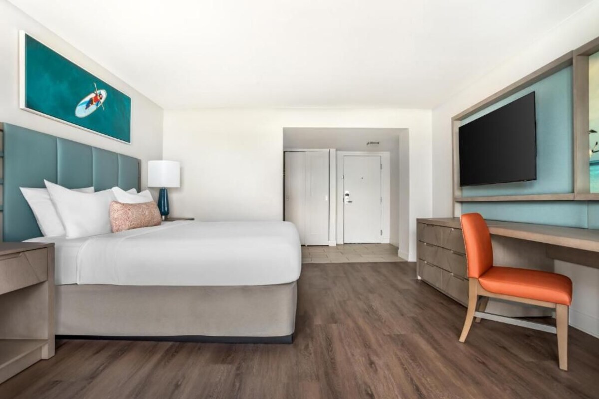 Una habitación de hotel con una cama, un escritorio, una silla, un televisor y un cuadro en la pared.