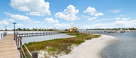 A wooden boardwalk leading to a beach with a small building and a dock extending into the water.