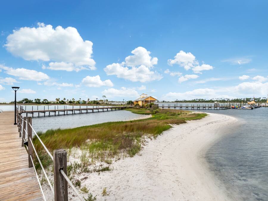 A wooden boardwalk leading to a beach with a small building and a dock extending into the water.