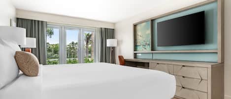 A hotel room with a large bed, a desk with a flat-screen TV, and a view of greenery outside.