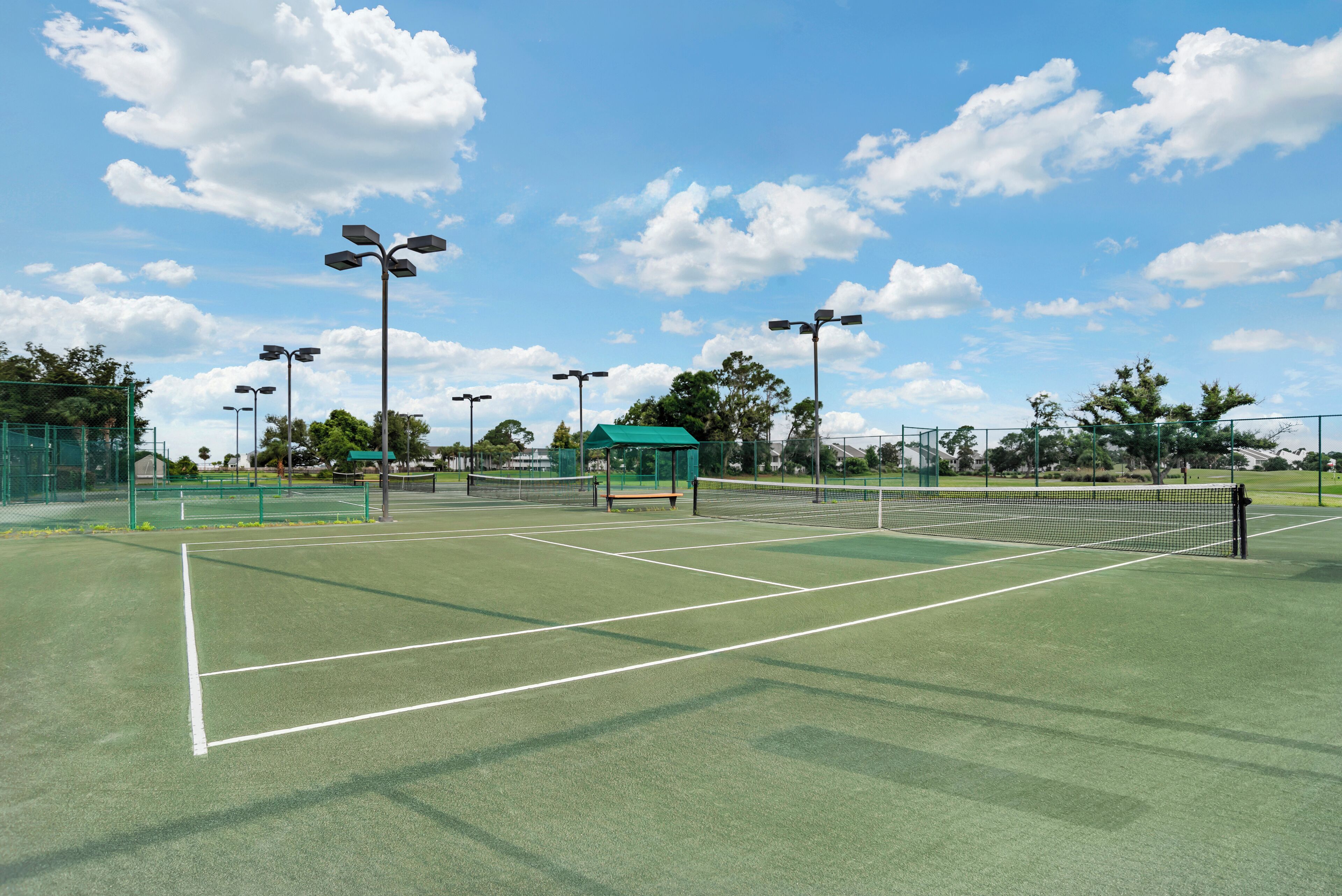 A tennis court with a net, surrounded by green fencing, and multiple tennis lights.