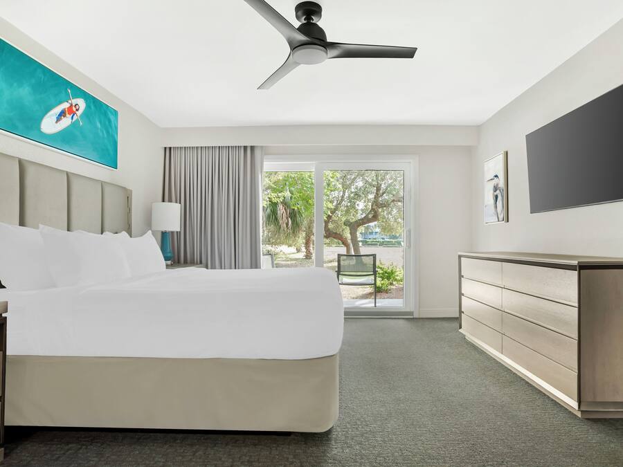 A hotel room with a bed, a desk, a chair, a TV, a ceiling fan, and a view of the outdoors.