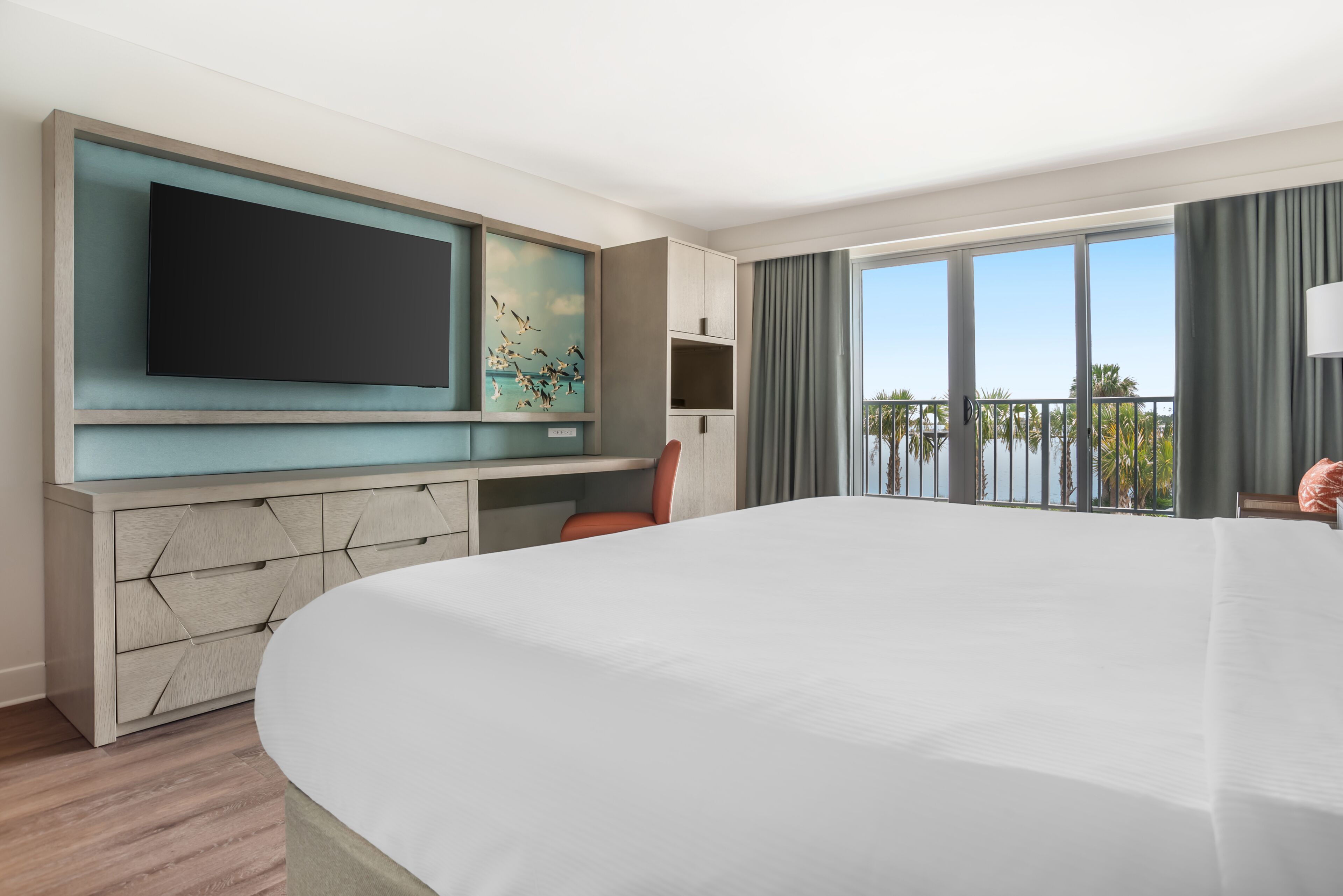 A hotel room with a large bed, a flat-screen TV, a desk with a chair, and a balcony with a view of the sea.