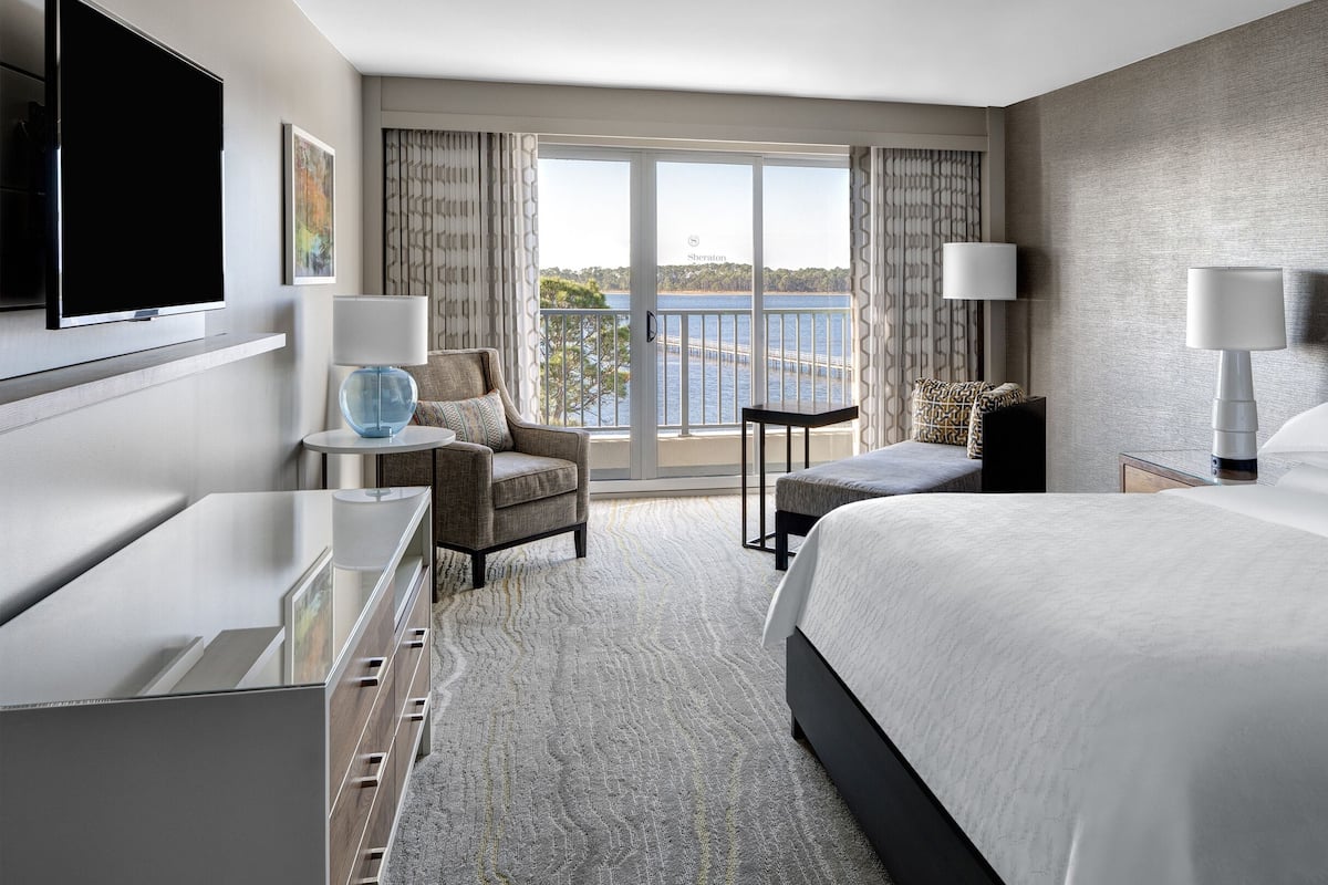 A hotel room with a large bed, a desk, a chair, and a view of the sea through the window.