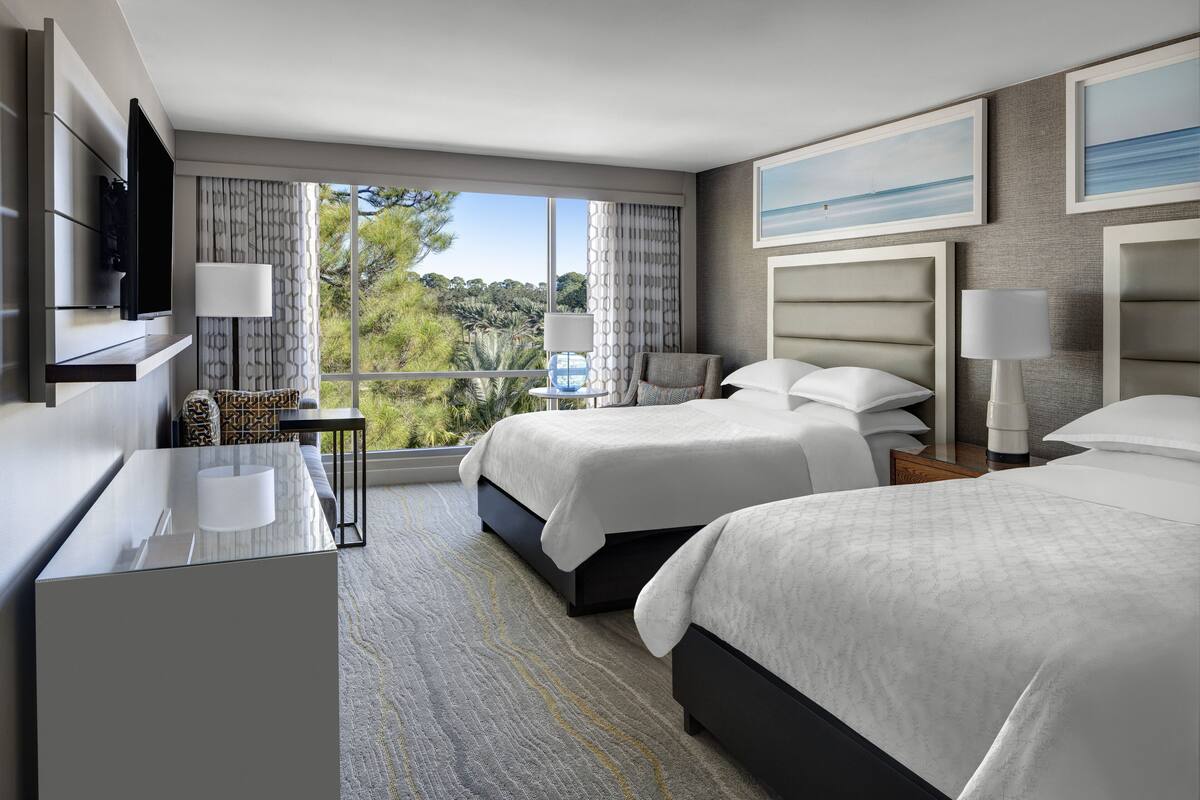 A hotel room with two beds, a desk, a TV, and a large window with a view of greenery.