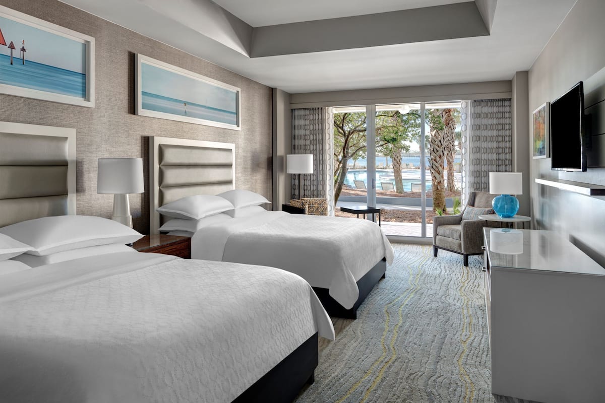 A hotel room with two beds, a TV, a desk, and a view of the outdoors.