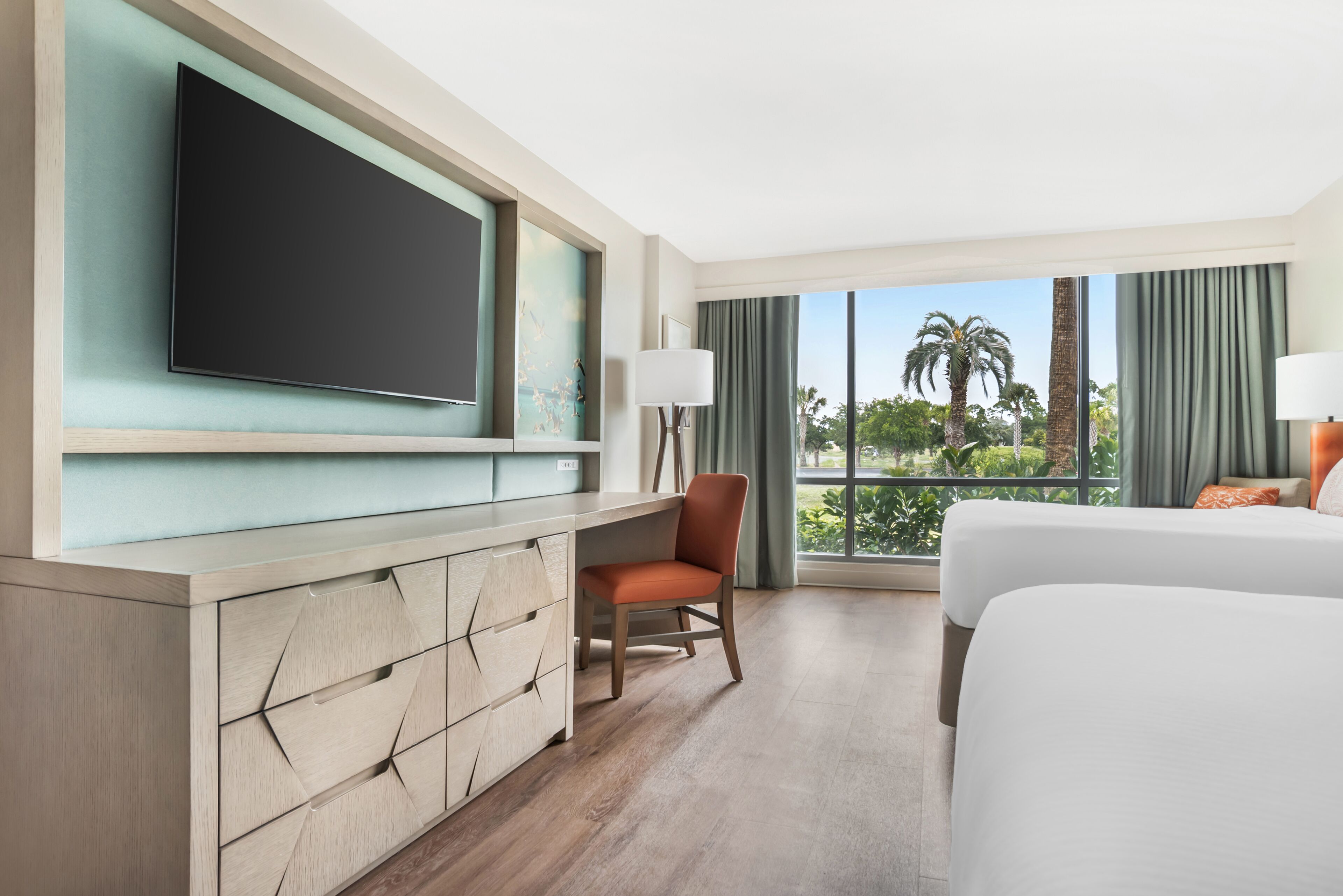 Hotelkamer met een grote flatscreen televisie, een bureau met lades, een stoel, een bed en uitzicht op palmbomen door het raam.