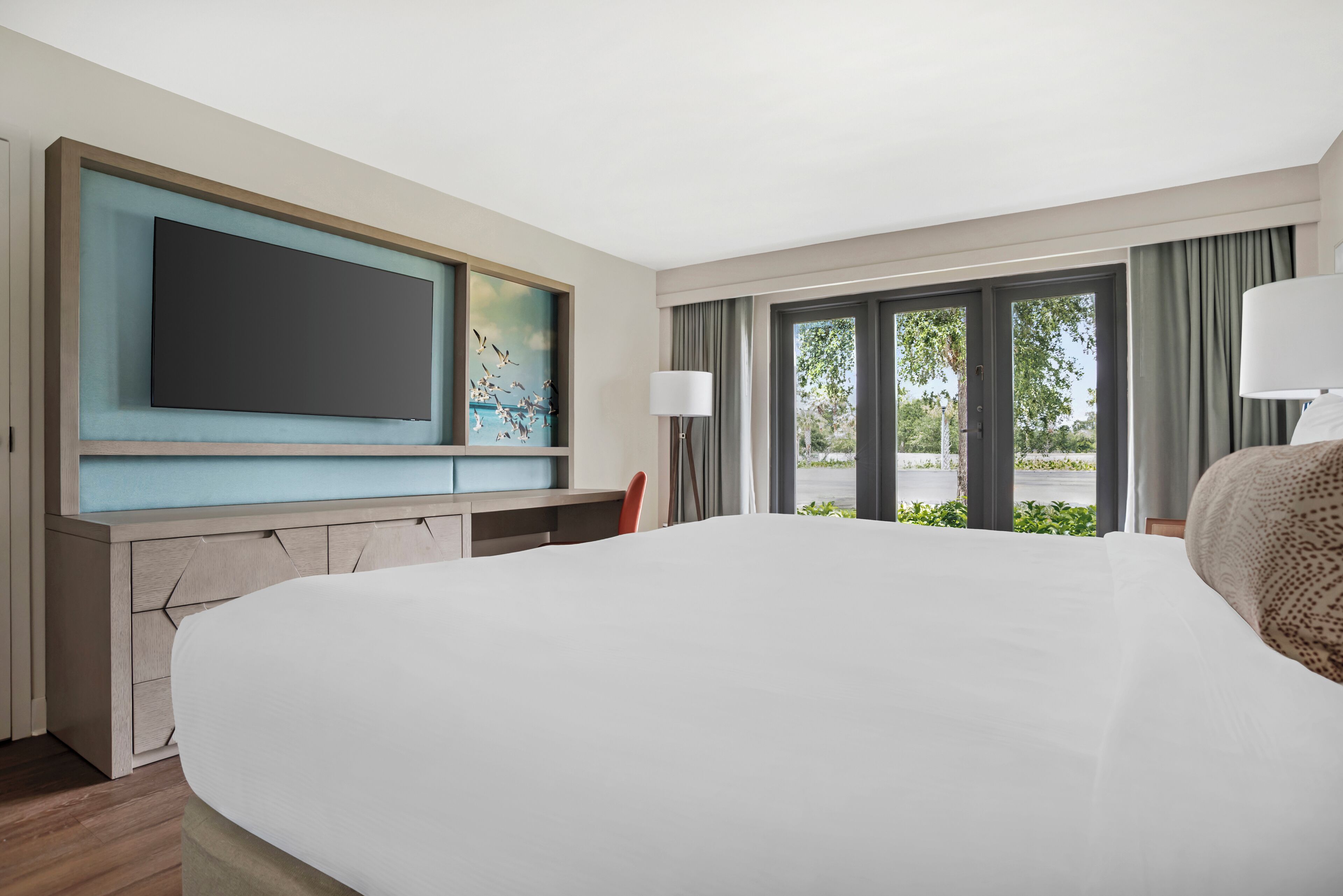 A hotel room with a large bed, a flat-screen TV mounted on the wall, a desk with a chair, and a window with curtains.