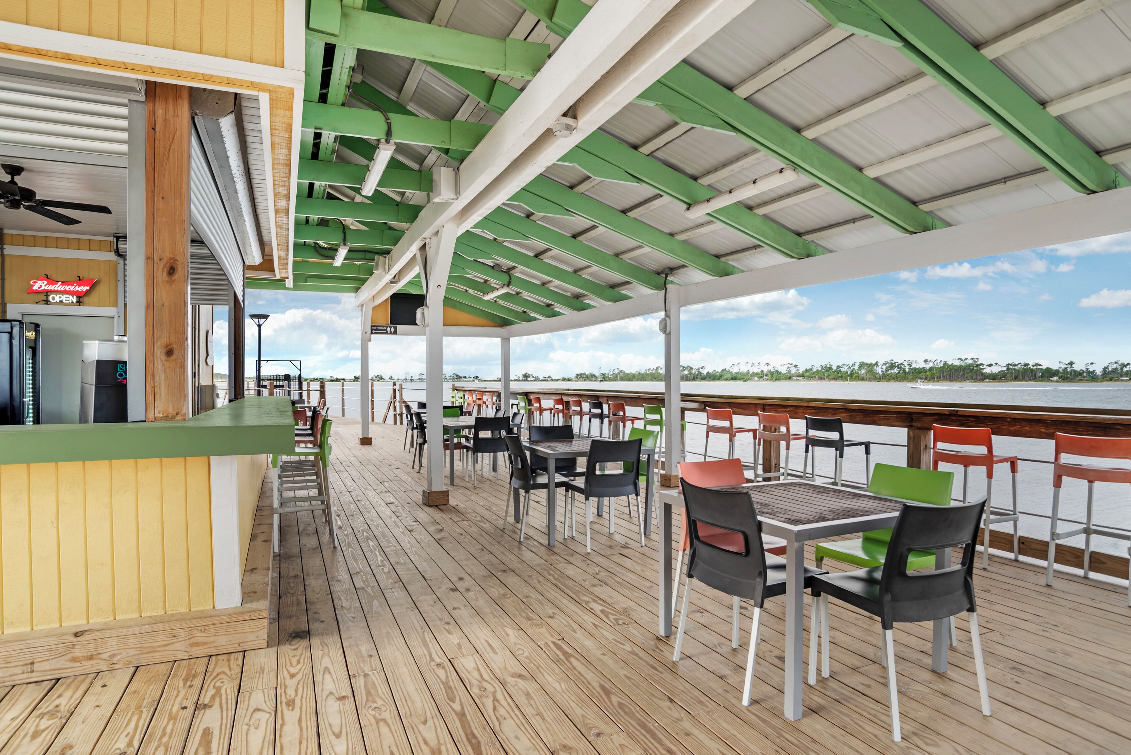 A covered outdoor deck with tables and chairs, a Budweiser sign, and a view of the water.