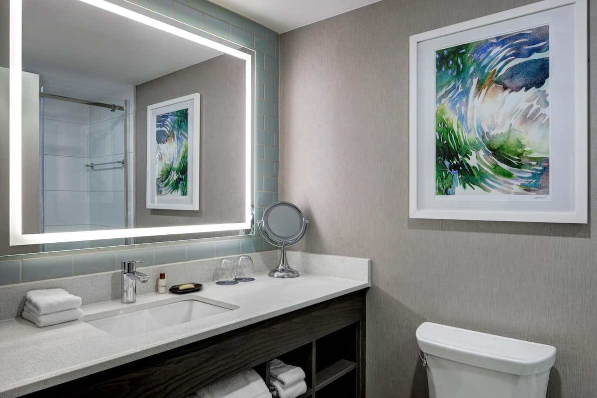 A modern bathroom with a large mirror, a sink, a toilet, and a framed painting on the wall.
