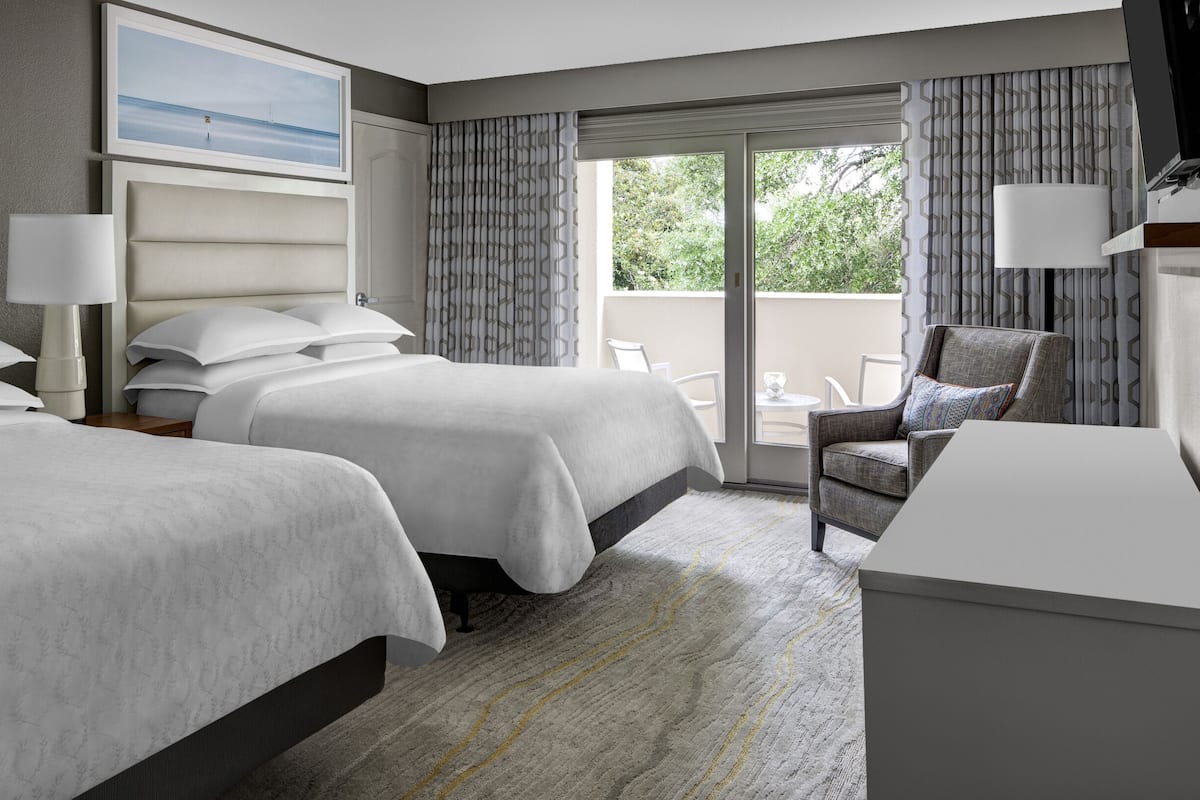 A hotel room with two beds, a desk, a chair, and a balcony with a view of greenery.