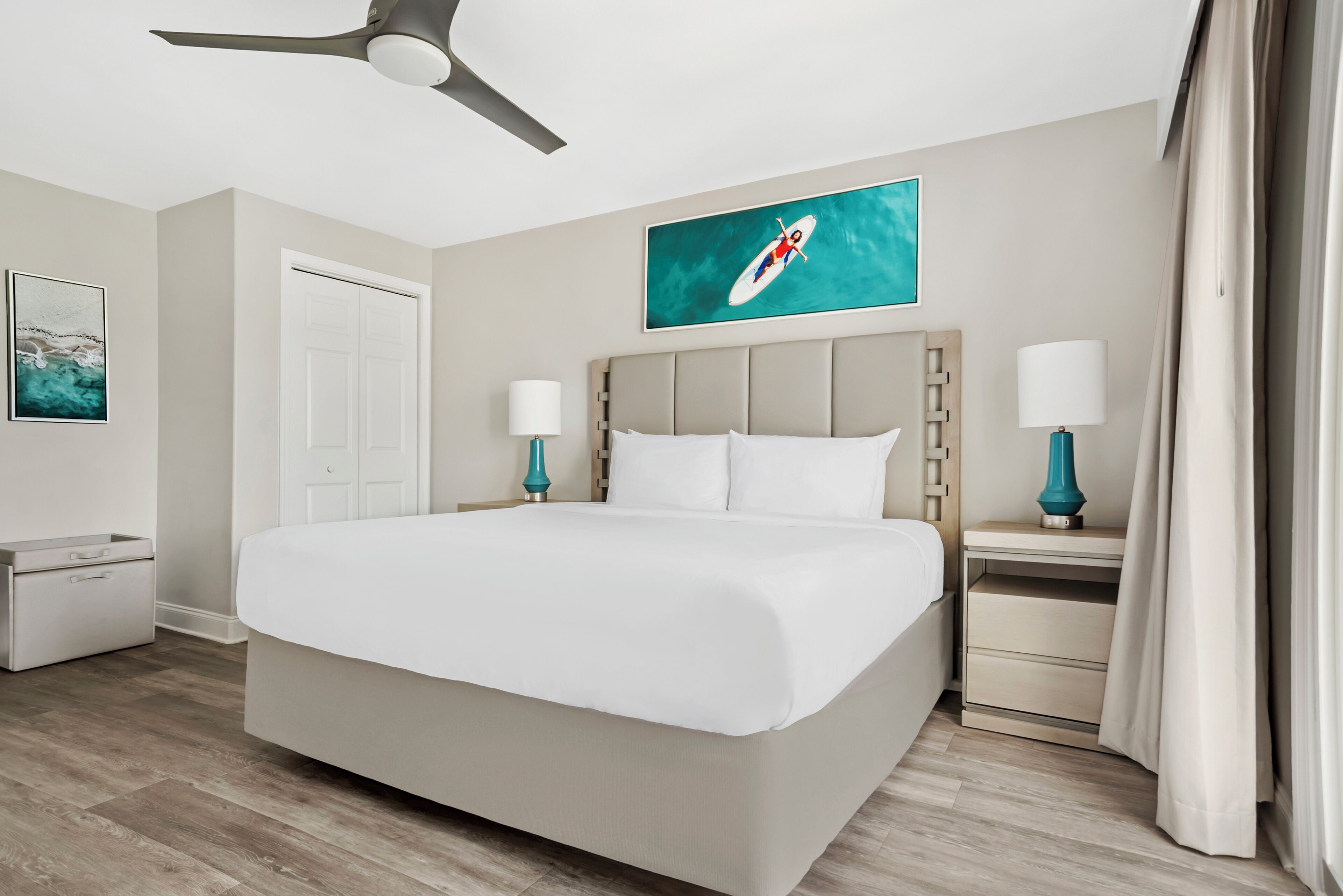 Habitación de hotel con una cama grande, ventilador de techo, mesita de noche con lámpara, una puerta y un cuadro en la pared.