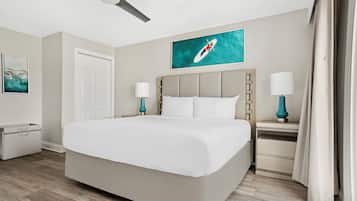 Een hotelkamer met een groot bed, een plafondventilator, een nachtkastje met een lamp, een deur en een schilderij aan de muur.