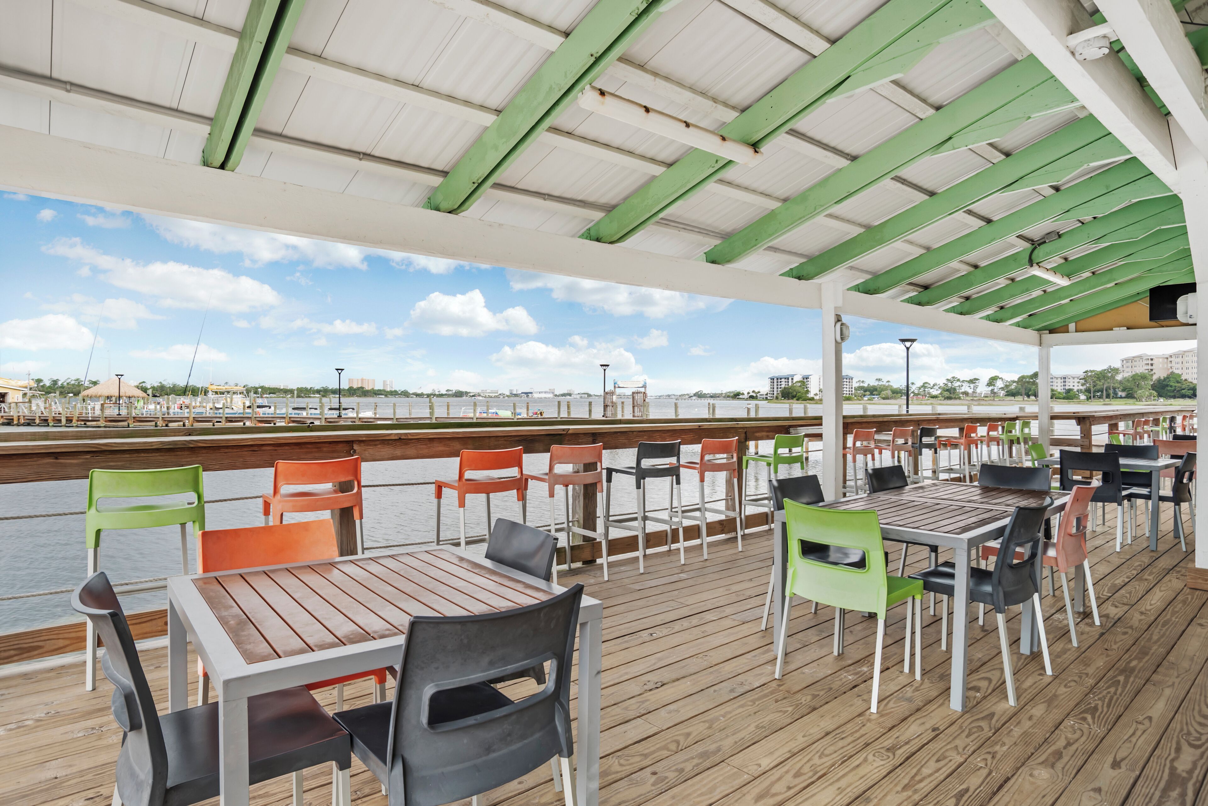An outdoor dining area with wooden tables and chairs, a view of the water, and a covered section with green beams.