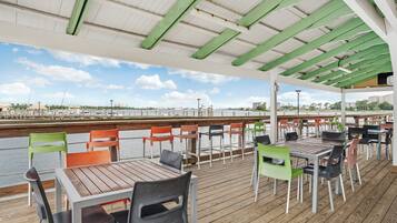 An outdoor dining area with wooden tables and chairs, a view of the water, and a covered section with green beams.
